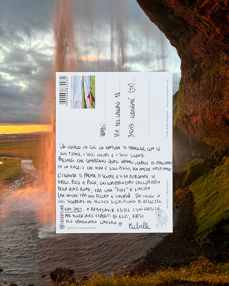 Cartolina manoscritta dall'Islanda tenuta in primo piano davanti alla cascata Seljalandsfoss illuminata dal tramonto.